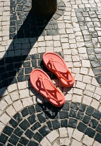 Tongs de couleur corail sur un pavement en mosaïque de pierres noires et blanches avec un arbre projetant une ombre à proximité.