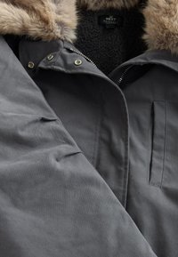 Parka gris foncé avec un col en fausse fourrure, équipée d'une fermeture éclair, de boutons-pression et d'une poche avant. Doublure intérieure douce et texturée visible.