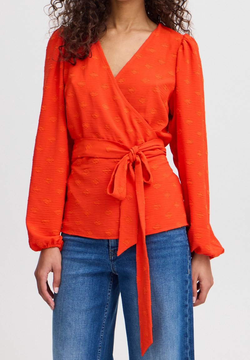 Femme portant un chemisier portefeuille rouge à manches longues avec un nœud à la taille et un jean bleu en denim sur un fond clair uni.