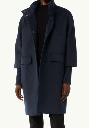 Donker marineblauwe wollen jas met hoge kraag, klepzakken en mouwlengte tot halverwege de onderarm, gedragen over een geribbelde donkere trui en zwarte broek.