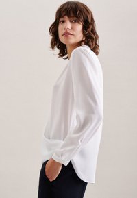 Weiße Bluse mit langen Ärmeln, Rundhalsausschnitt, weichem Stoff, lässiger Passform und dezentem Fall. Verfügt über umgeschlagene Bündchen und Seitenschlitze.