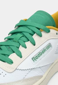 Tenisky Reebok s bielym koženým zvrškom, zelenými šnúrkami, žltými detailmi a textúrovanou bočnou časťou s logom. Viditeľné odpruženie.
