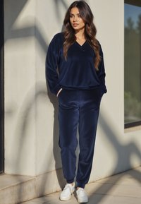 Tuta in velluto morbido di colore blu navy, con maglia a scollo a V e pantaloni con vita elasticizzata e tasche, abbinati a sneaker bianche.