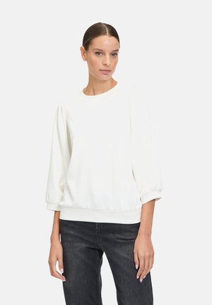 Weißer, leichter Pullover mit Rundhalsausschnitt, puffärmeln und gerafftem Saum. Kombiniert mit dunklen, strukturierten Jeans.