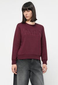Una mujer con el cabello lacio y oscuro lleva una sudadera burdeos con "PARIS" en relieve, combinada con pantalones vaqueros negros desgastados y holgados, de pie contra un fondo blanco.
