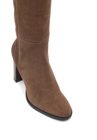 Bota de ante marrón, de caña alta, con punta afilada y tacón tapered. Detalles de costura en el eje y suela negra lisa.