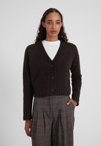Gilet en tricot marron avec un profond décolleté en V, un ourlet côtelé et quatre boutons noirs. Porté sur un T-shirt blanc et un pantalon à carreaux.
