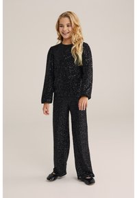 Musta paljeteilla varustettu urheilupuku, jossa on pitkähihainen paita ja löysät housut. Sujuva pinta ja hohtava viimeistely, yhdistettynä mustiin kenkiin.