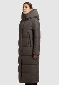 Lange pufferjas in donkergrijs, met een hoge kraag, een ritssluitingzak opzij en een gewatteerd patroon. Glad oppervlak en warme uitstraling.