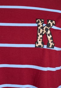 Czerwona koszula z krótkim rękawem z niebieskimi paskami i aplikacją w kształcie litery "K" w zwierzęcy wzór na lewej stronie. Wykonana z miękkiego materiału bawełnianego.