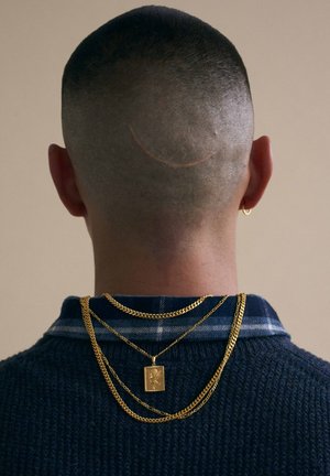 Short gold chains layered over a navy textured sweater, with a subtle blue plaid collar. A smile-shaped line is visible on the shaved head.