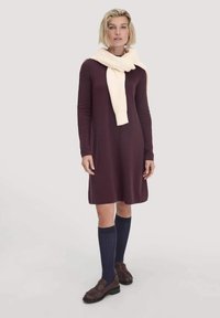 Frau in einem langärmeligen, pflaumenfarbenen Kleid, mit einem cremefarbenen Schal um den Hals, marineblauen knielangen Socken und braunen Mokassins, die vor einem schlichten Hintergrund steht.