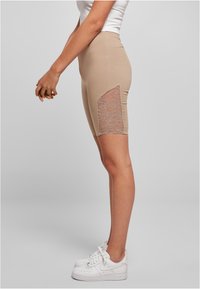 Beige fietsbroek met een kantpaneel aan de zijkant, gecombineerd met witte sneakers. Glad materiaal met een aangesloten design, waarin het kantdetail goed tot zijn recht komt.