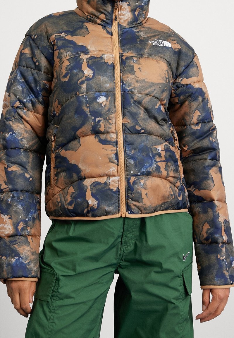Polstret jakke i brun, blå og sandfarvet med camouflage-mønster. Har høj krave og lynlås, parret med grønne cargo-bukser.