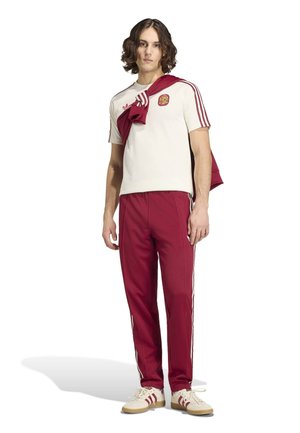 Jeune homme portant un maillot de football blanc de l’Espagne avec des rayures bordeaux et un pantalon bordeaux, une veste bordeaux posée sur ses épaules.