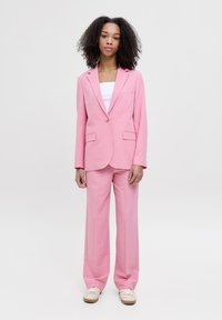 Roze blazer en bijpassende wijde broek van gladde stof, met een enkele knoopsluiting en voorzakken. Vergezeld door een witte top.
