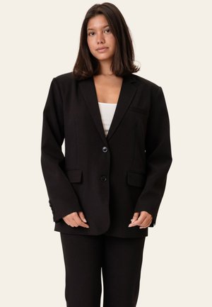 Junge Frau mit glatten dunklen Haaren, die einen übergroßen schwarzen Blazer mit zwei Knöpfen und passende schwarze Hosen über einem weißen Oberteil trägt.