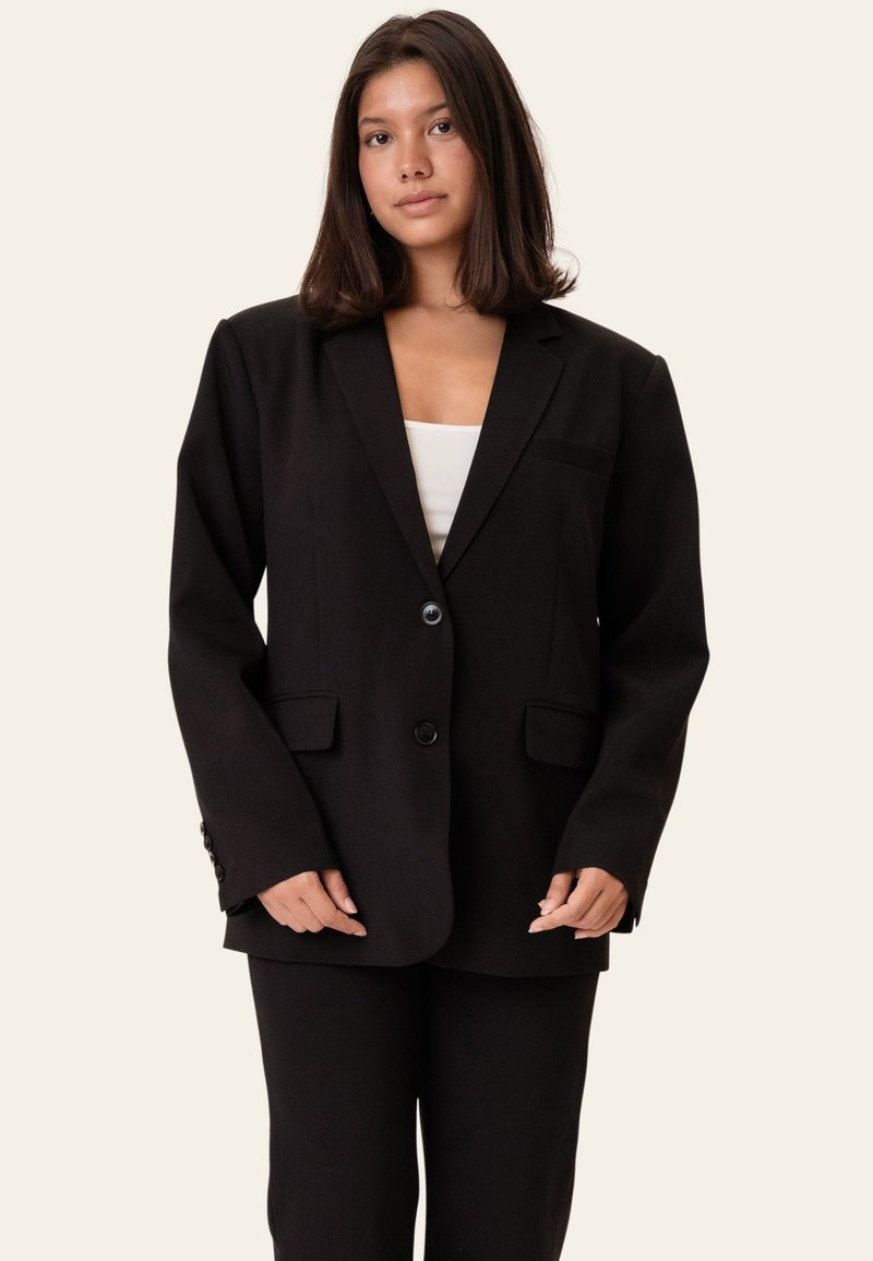 Jeune femme aux cheveux raides et foncés portant un blazer noir surdimensionné à deux boutons et un pantalon noir assorti par-dessus un haut blanc.