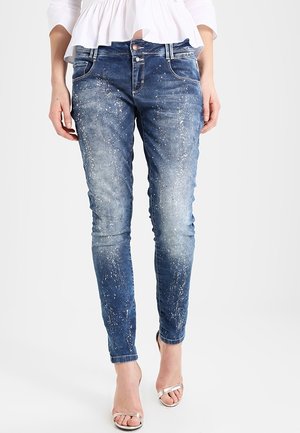 Jean bleu slim avec des éclaboussures de peinture blanche, porté avec des talons ouverts argentés et un chemisier blanc.