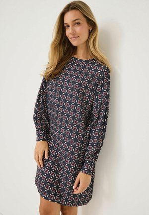 Kvinna som står mot en vit vägg iförd en långärmad svart klänning med röda och vita geometriska stjärnmönster.