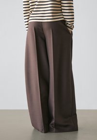 Persona che indossa pantaloni a vita alta a gamba larga con pieghe frontali in due tonalità di marrone, in piedi su un pavimento semplice contro una parete chiara.