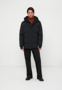Chaqueta acolchada negra con capucha, con patrones acolchados rectangulares y un bolsillo frontal. Llevada sobre un suéter de cuello alto marrón y pantalones negros.
