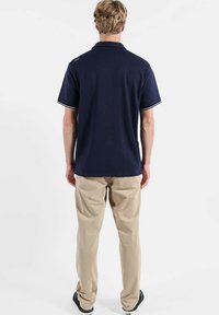 Polo bleu marine avec accents blancs, manches courtes et col classique, associé à un pantalon beige et des baskets noires.