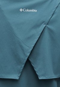 Ύφασμα σε τιρκουάζ με σχεδιασμό επικαλυπτόμενων πτερυγίων και λευκό λογότυπο Columbia κεντημένο κοντά στο επάνω κέντρο.