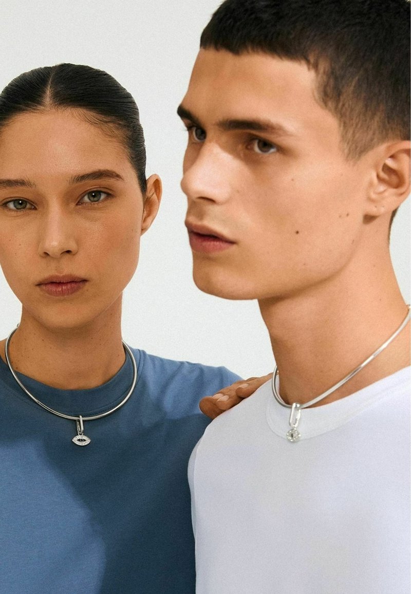 Silverhalsband i form av choker som bärs av två personer; en har ett runt hänge, medan den andra har en design med ett öga. Texturerade t-shirts i blått och vitt.