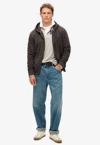 Sudadera gris con cremallera sobre un polo gris, jeans de tiro alto azules y zapatillas blancas con detalles marrones. Ajuste casual y relajado.