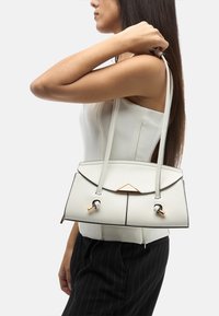 Borsa a tracolla bianca in pelle con design strutturato, dettagli neri e una decorazione triangolare dorata sul cappuccio. Due lunghe cinghie per il trasporto.