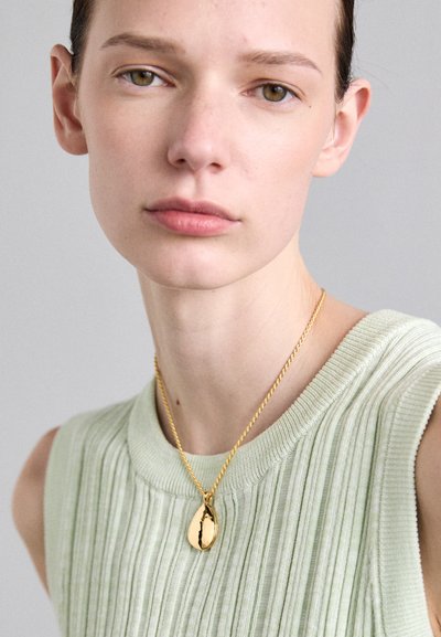 Gold pendant necklace on a thin chain worn over a light green textured sweater with a ribbed design. The background is neutral gray.