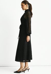 Black lace dress with long, puffed sleeves, a high neckline, and a tied waist detail. Skirt reaches mid-calf. Worn with black heeled slip-on shoes.