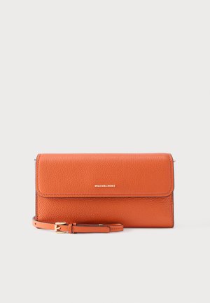 Oranža raupja ādas rokassomu ar zeltainu Michael Kors logotipu un saskaņotu regulējamu siksnu ar zelta sprādzi uz pelēka fona.