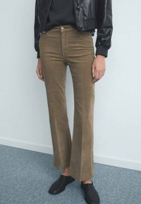 Pantaloni a zampa in velluto marrone, vita media, texture liscia, con quattro tasche e chiusura con bottone, abbinati a una giacca e scarpe in pelle nera.