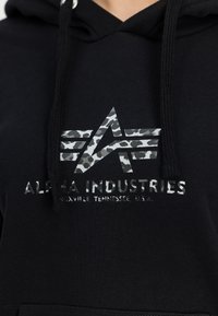Sudadera negra con un logotipo de estrella de camuflaje reflectante y el texto "ALPHA INDUSTRIES, KNOXVILLE, TENNESSEE, U.S.A." en color plateado.