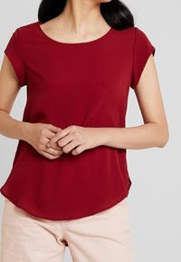 Bordowa bluzka z krótkim rękawem, z okrągłym dekoltem, gładką fakturą i zaokrąglonym dołem, zestawiona z jasnoróżowymi spodniami.