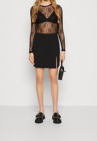 Svart spetsblus med långa ärmar, över en svart figurnära kjol med sidov slit, tillsammans med svarta kraftiga loafers och en liten handväska.