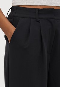 Black trousers with a smooth texture, featuring pleats, a waistband, and side pockets, showcased on a partially visible hand.