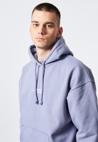 Ljuslila hoodie med känguruficka, dragsko i huvan och vit textlogotyp på bröstet. Mjuk, texturerad tyg.