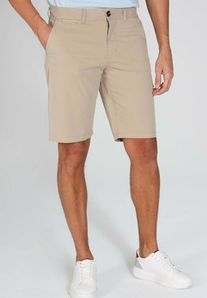 Homme portant un short beige mi-genou et des baskets blanches, debout avec une main dans la poche devant un fond uni.
