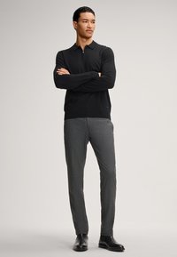 Schwarzer gepolsterter Polo-Pullover mit langen Ärmeln, kombiniert mit grauen strukturierten Hosen und schwarzen Anzugschuhen. Minimale Details, taillierte Form.