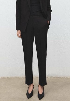 Personne portant un pantalon noir taille haute sur mesure, un blazer noir et des talons noirs à bouts pointus, debout sur un sol neutre devant un mur blanc.