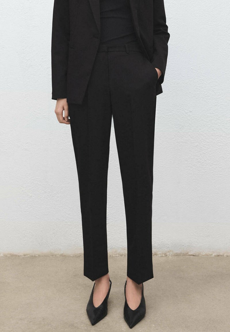 Personne portant un pantalon noir taille haute sur mesure, un blazer noir et des talons noirs à bouts pointus, debout sur un sol neutre devant un mur blanc.