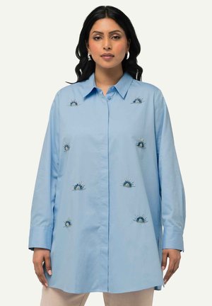 Camicia azzurro chiaro a maniche lunghe con colletto a bottoni, caratterizzata da intricati motivi ricamati blu che ricordano i raggi del sole sul davanti.