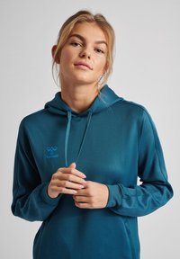 Sweatshirt à capuche en tissu teal avec une texture lisse. Présente un logo sur le devant et des cordons de serrage, avec des poignets côtelés et une coupe décontractée.