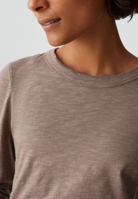 Camicia marrone a maniche lunghe realizzata in morbido tessuto, con collo tondo e una sottile trama. Sono visibili i dettagli delle cuciture lungo il taglio del collo.
