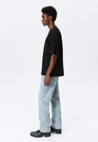 Ung mann står på siden iført en svart oversized t-skjorte, lyseblå jeans og svarte støvler mot en hvit bakgrunn.
