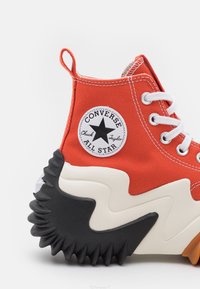 Sneaker alta in tela rossa con lacci bianchi, dotata di una suola in gomma nera con un motivo a zigzag e una patch circolare con logo sul lato.