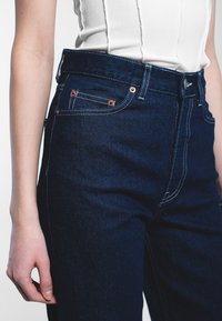 Tmavé džíny s vysokým pasem, s měděnými kovovými prvky, kontrastním bílým prošíváním a klasickým pětikapsovým designem.